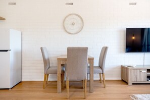 A modern kitchen with a white refrigerator, a dining table with four chairs, and a wall-mounted clock. - Stirling Breeze – One Bedroom Apartment Near Beach & Transport (Mosman Park)