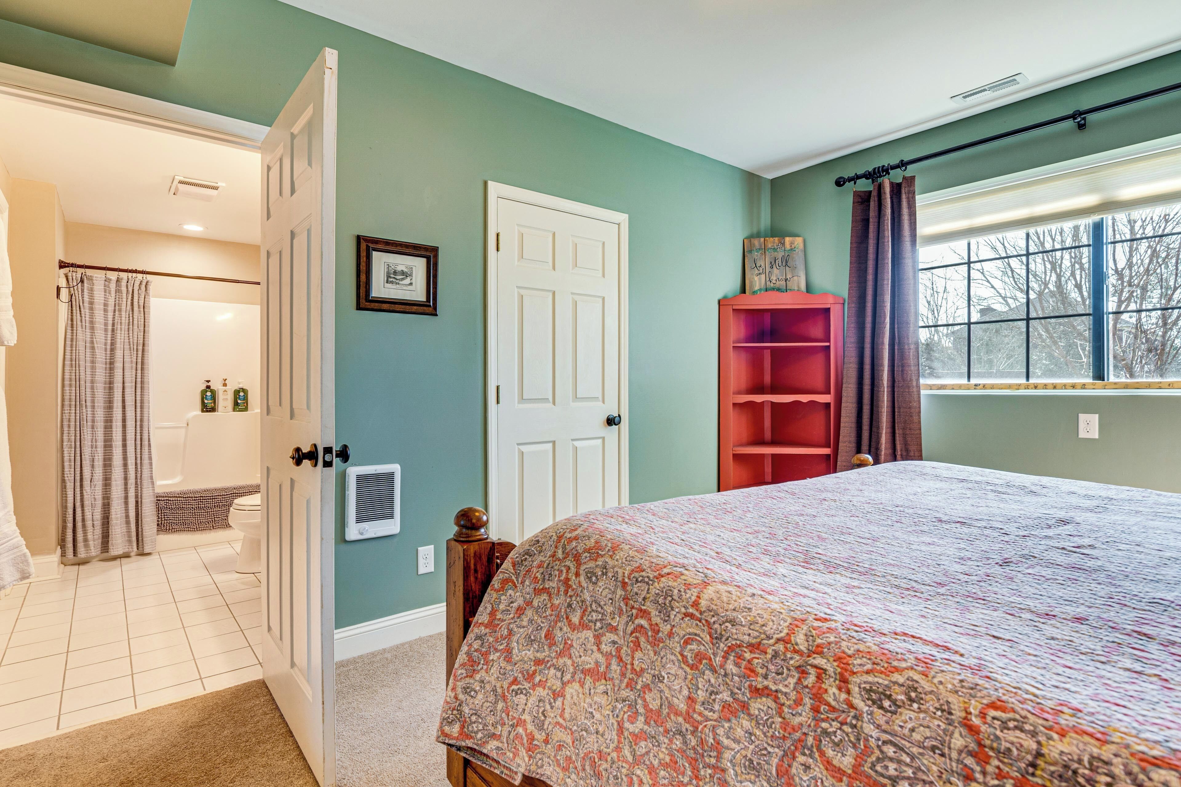 A bedroom with a bed, a door leading to a bathroom, a window with curtains, and a red shelf.