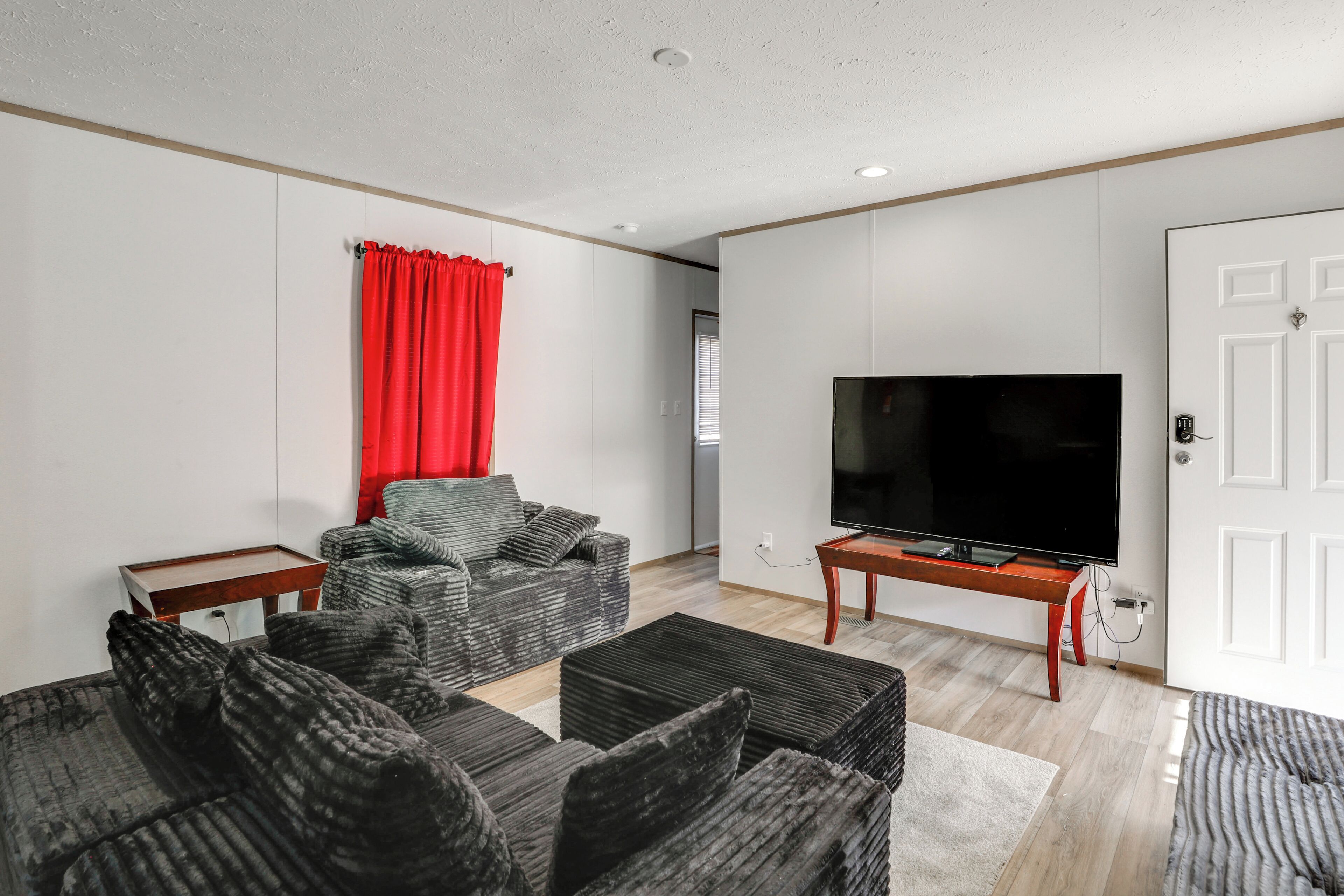 Een moderne woonkamer met een grote flatscreen televisie, grijze gestoffeerde banken, een rood gordijn en een witte deur.