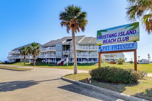 A beachfront hotel named Mustang Island Beach Club, featuring a pet-friendly sign, multiple balconies, and a well-maintained lawn with palm trees. - Beach Getaway on Mustang Island: Pet Friendly! (Port Aransas)