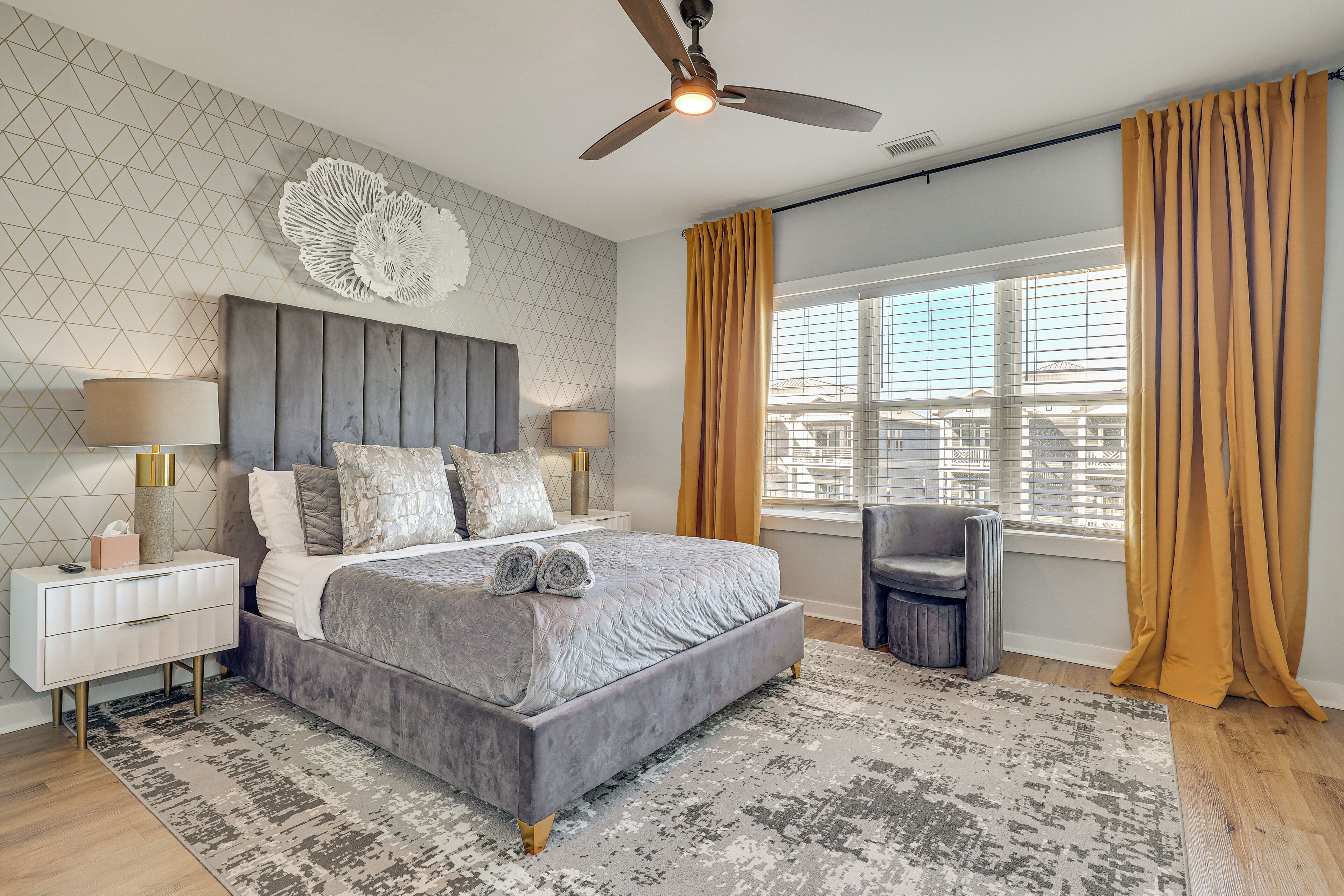 A modern bedroom with a large bed, a ceiling fan, a nightstand, a chair, and a window with curtains.