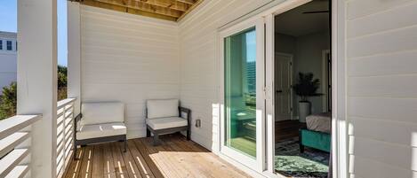 A covered deck with wooden flooring, white walls, and a sliding glass door leading to an interior space with a green sofa and a white chair.