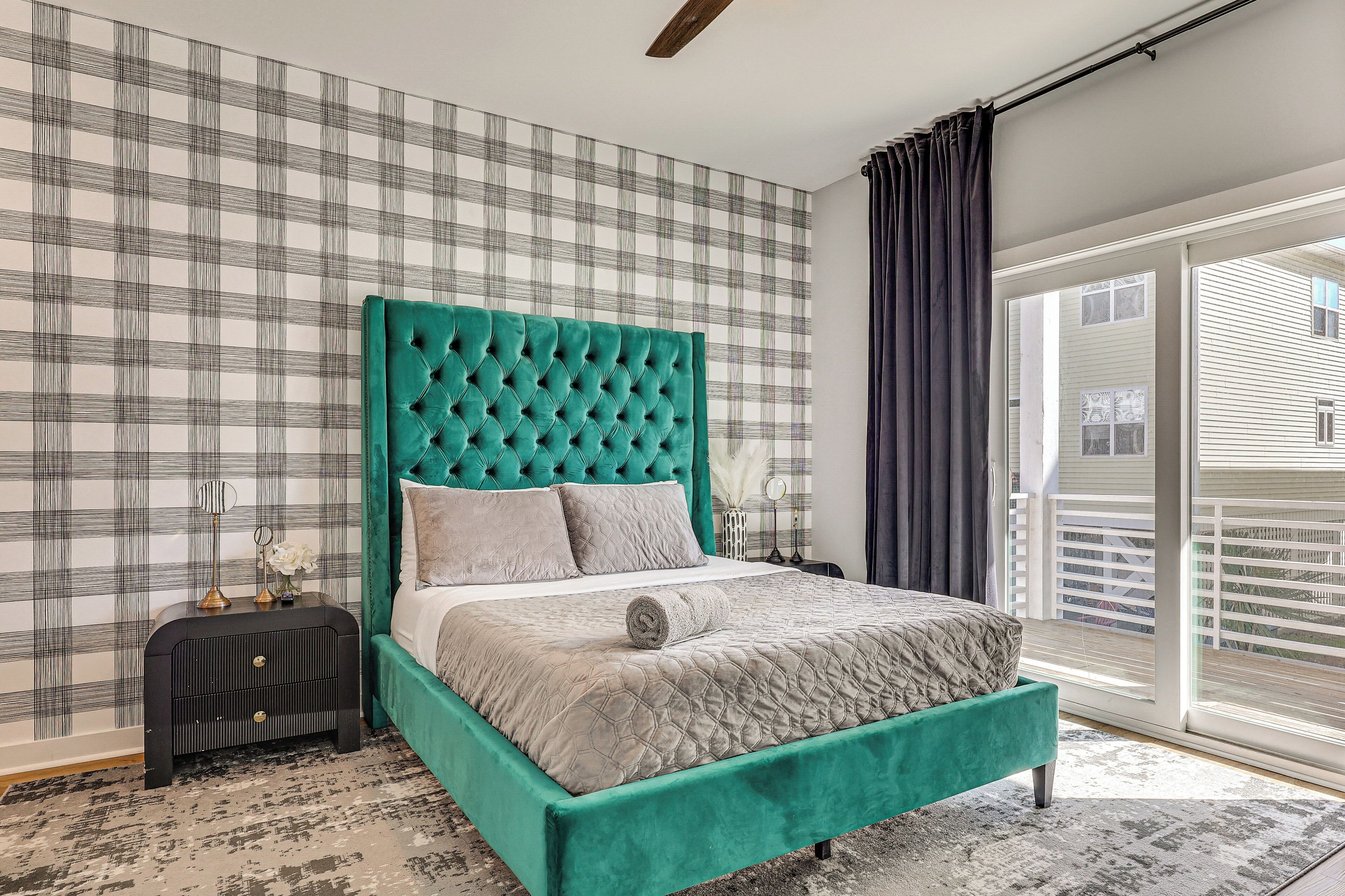 A bedroom with a checkered wallpaper, a tufted teal headboard, a bed with a quilted grey bedspread, and a nightstand with a lamp.