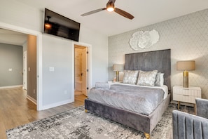 A bedroom with a bed, a ceiling fan, a TV, a decorative wall piece, and a nightstand with lamps. - Sun, Surf & Group Fun in Carolina Beach! Luxe Home (Carolina Beach)