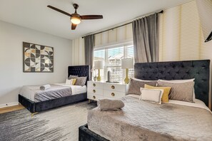 A bedroom with two beds, a ceiling fan, a window with blinds, a bedside table with lamps, and a wall art piece. - Sun, Surf & Group Fun in Carolina Beach! Luxe Home (Carolina Beach)