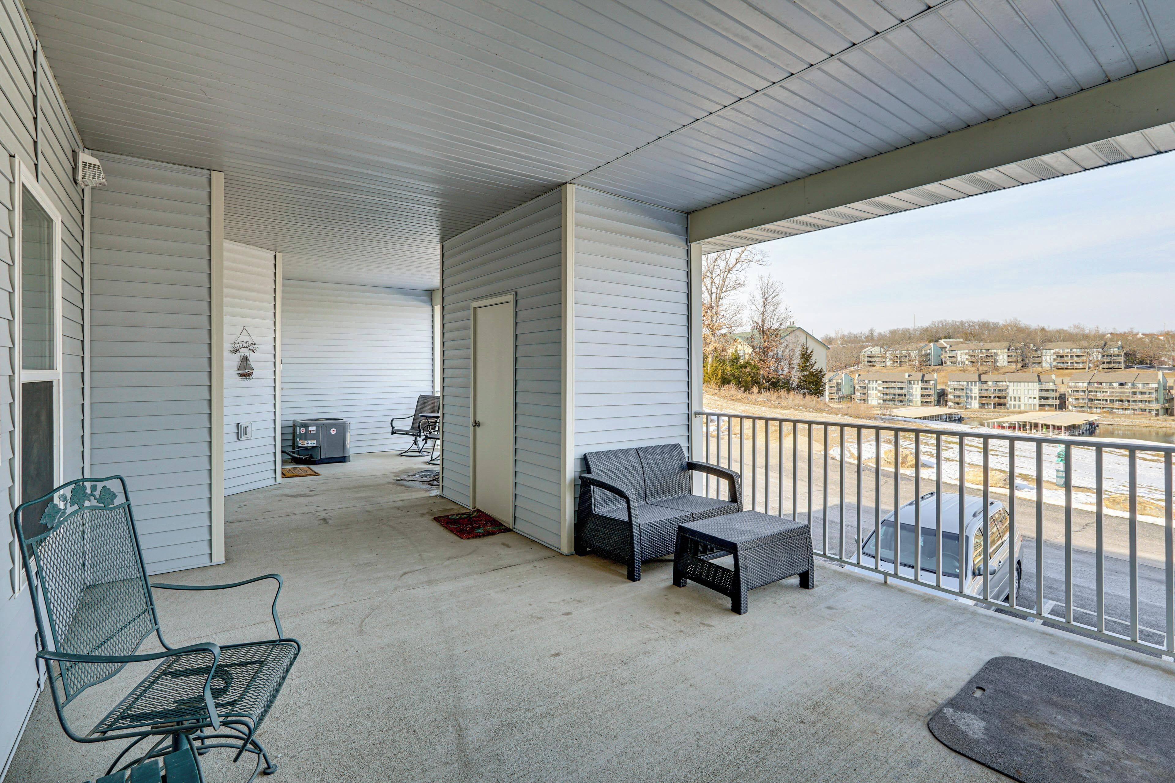 A covered porch with a seating area, a grill, and a view of a parking lot and buildings in the background.