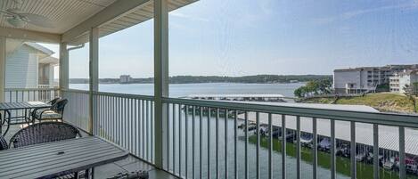 A covered balcony with a view of a body of water, a parking area, and buildings in the distance.