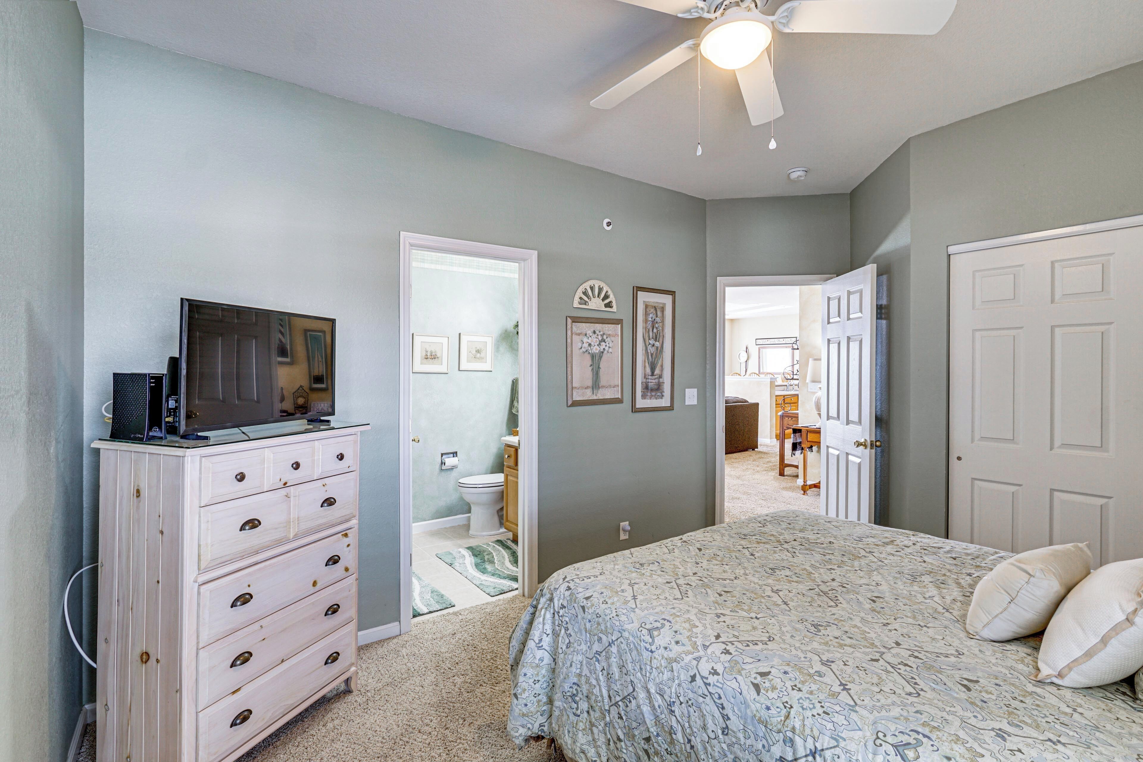A bedroom with a bed, dresser, ceiling fan, and a door leading to another room.