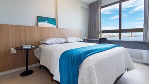 Quarto de hotel com cama, uma pequena mesa, um quadro e uma janela grande com vista para a paisagem urbana.