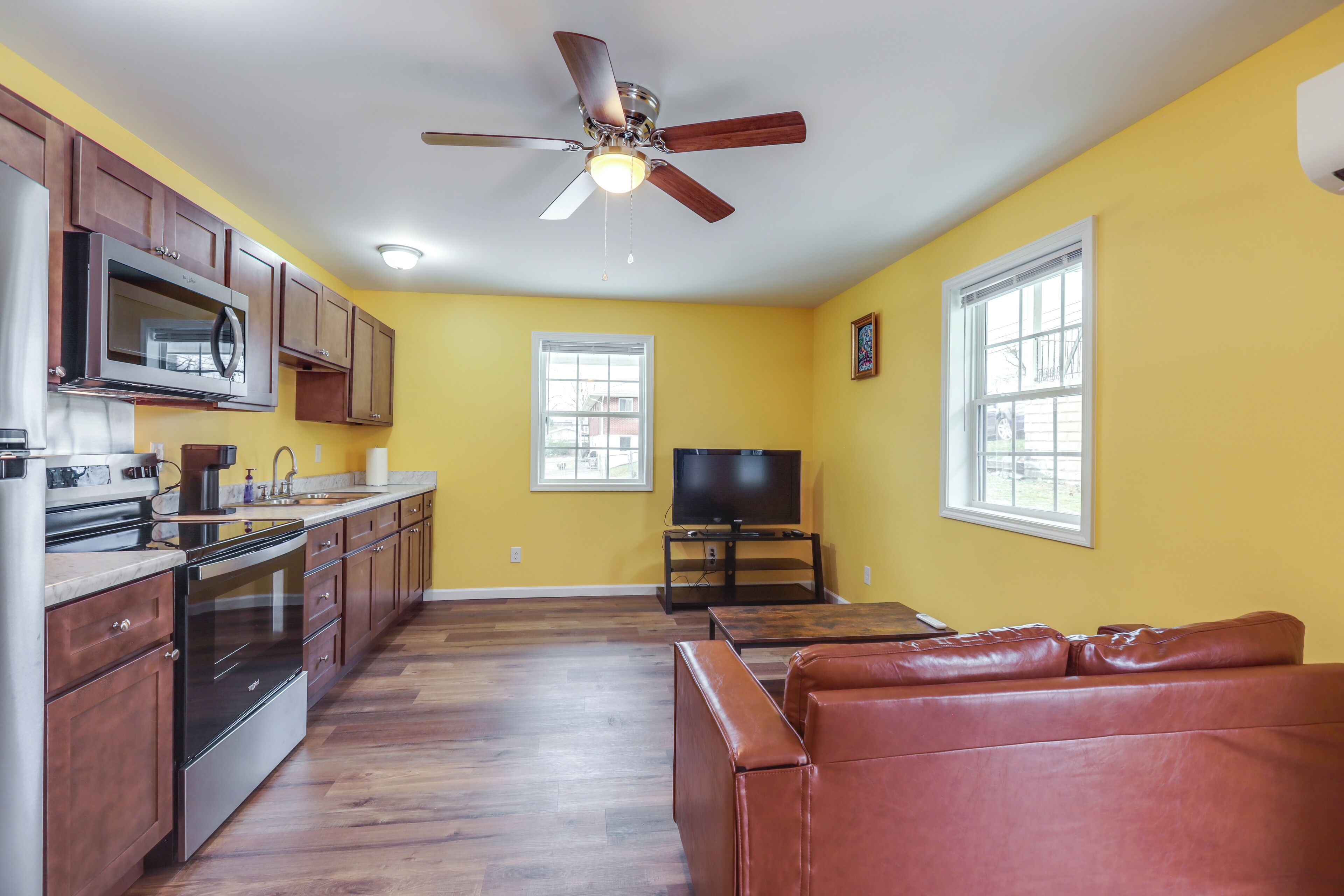 A kitchen with wooden cabinets, a microwave, and a stove. There is a brown leather couch and a television in the living area.