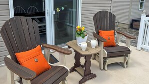 Twee Adirondack stoelen met oranje kussens, een klein tafeltje met een witte mok en een bloempot, en een schuifdeur met glas.