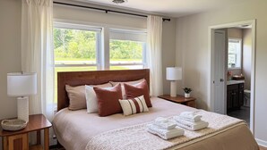Een net opgemaakt bed met hoofdbord, nachtlampjes en een raam met gordijnen.