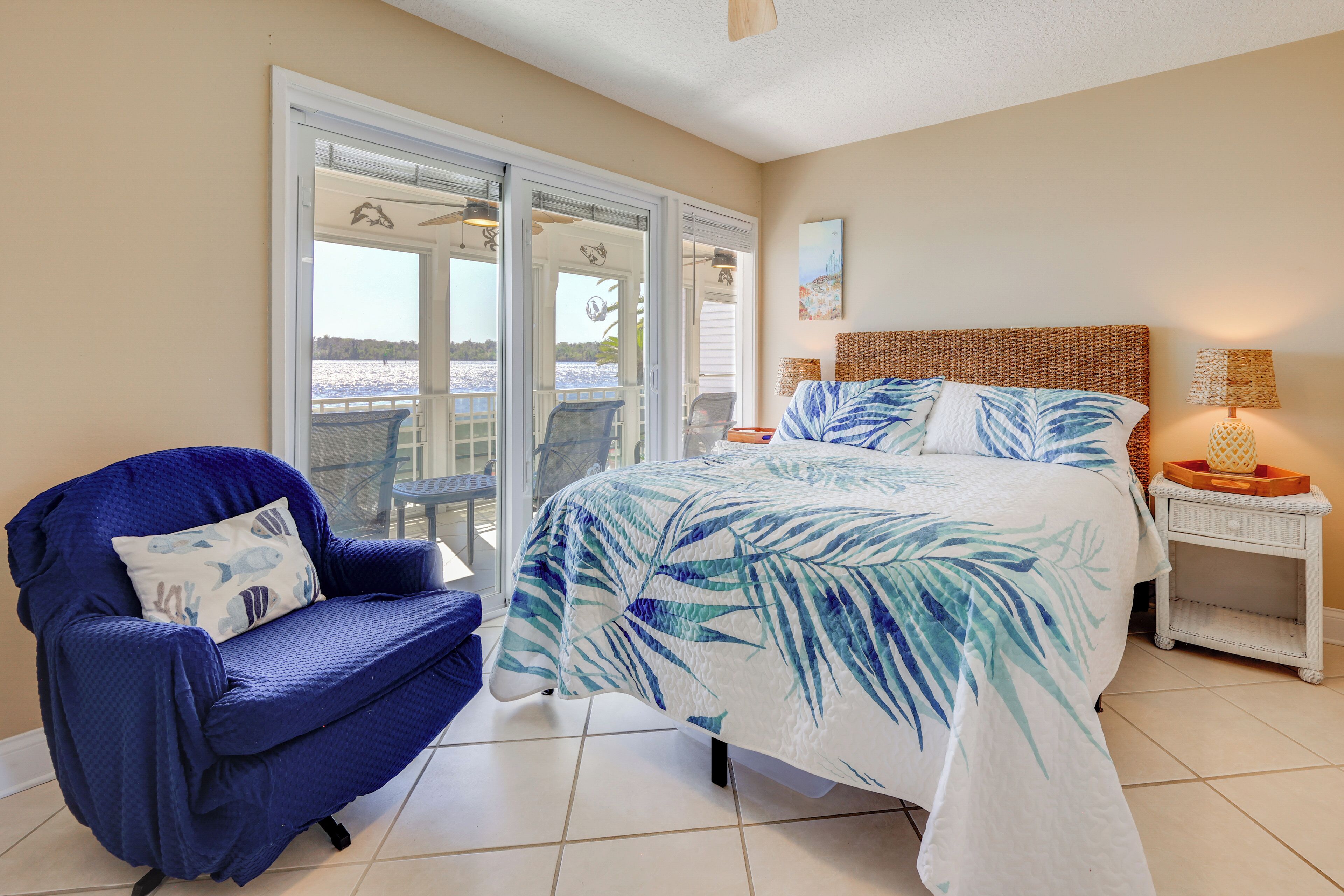 A bedroom with a bed, a blue armchair, a small table with a lamp, and a view through a sliding glass door.