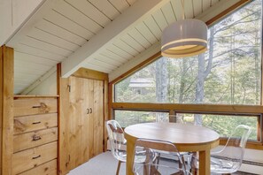 A wooden interior with a round table and chairs, a ceiling with wooden beams, and a large window with a view of trees. - Resort-style Community! Quechee Chalet w/ Deck (Quechee)