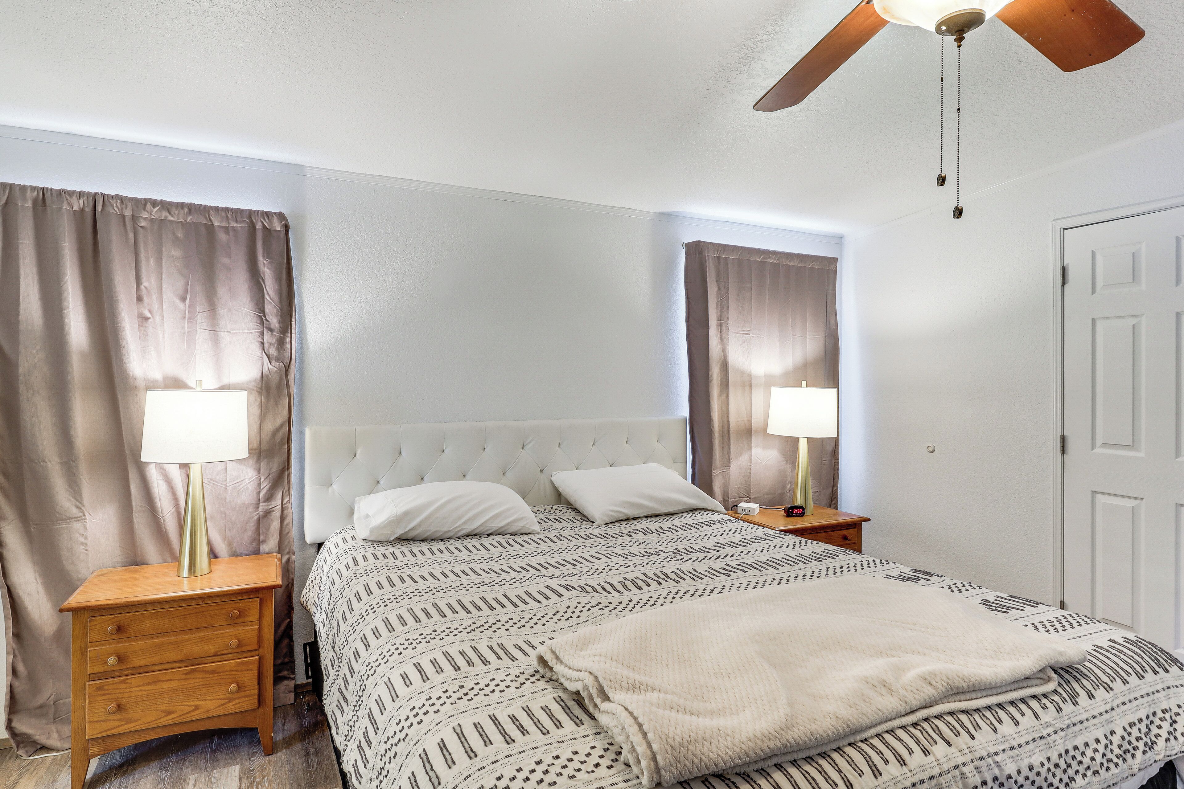Een net opgemaakt bed met een bedsprei met patroon, twee bedlampen, een houten nachtkastje met lades, en een plafondventilator.