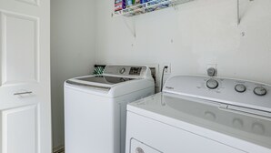 A laundry room with a front-loading washer and dryer, a shelf with cleaning supplies, and a white door. - Lake-view Townhome w/ Pool Access in Houston Area! (Missouri City)