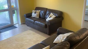 A living room with a brown leather sofa, a smaller sofa, and a glass door leading outside. - 2 Bed Cottage - Sleeps 5 - Pets - Parking - Garden (Liskeard)