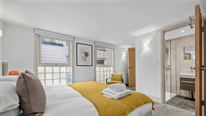 A hotel room with a bed, a yellow throw blanket, a brown pillow, a chair, a bathroom with a mirror, and a view of buildings through the windows. - Elegant Soho 1-Bed - Balcony, 2 Baths, Sleeps 4 (London)