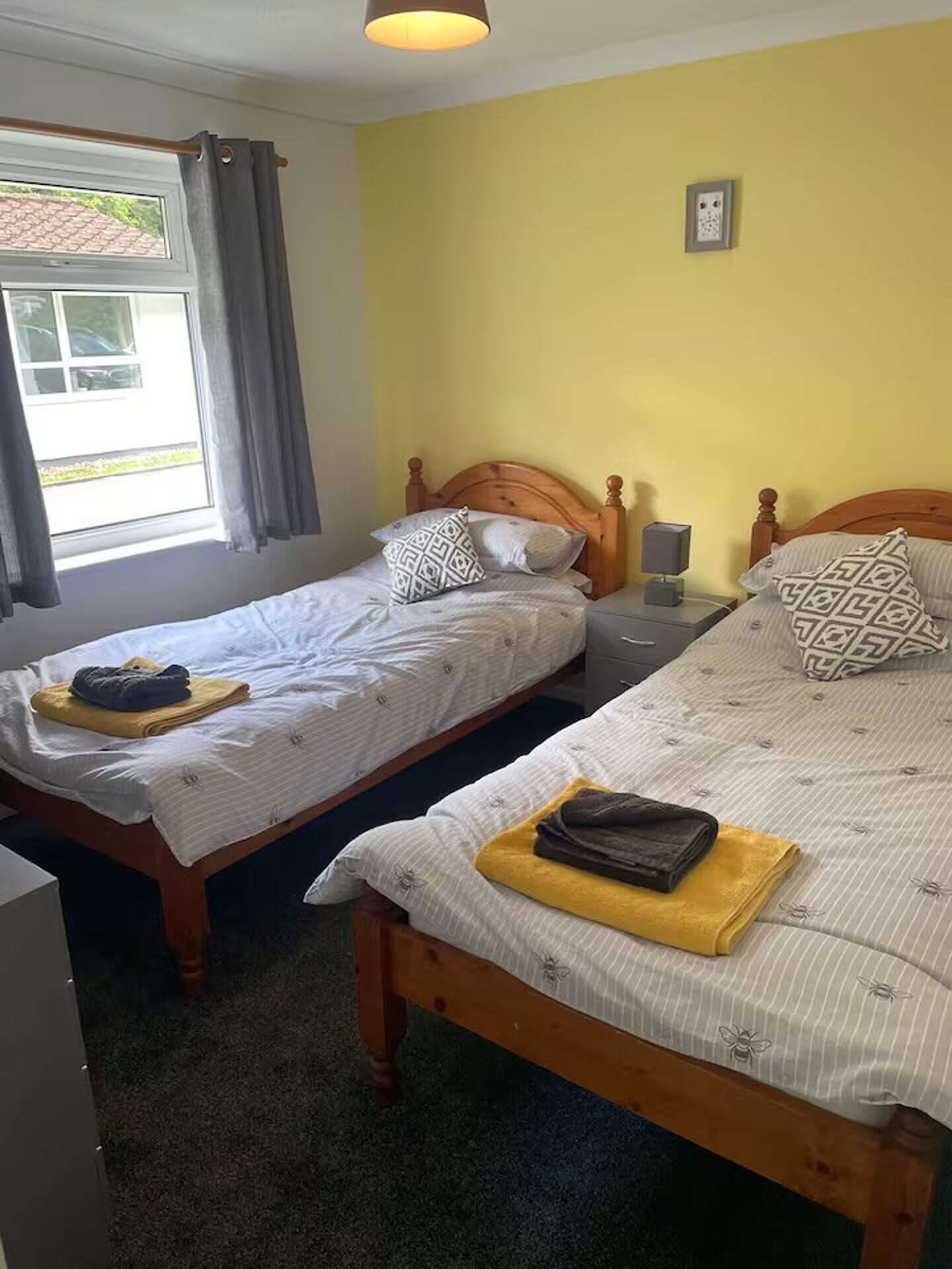 A room with two single beds, a window with curtains, a bedside table with a lamp, and a small picture on the wall.