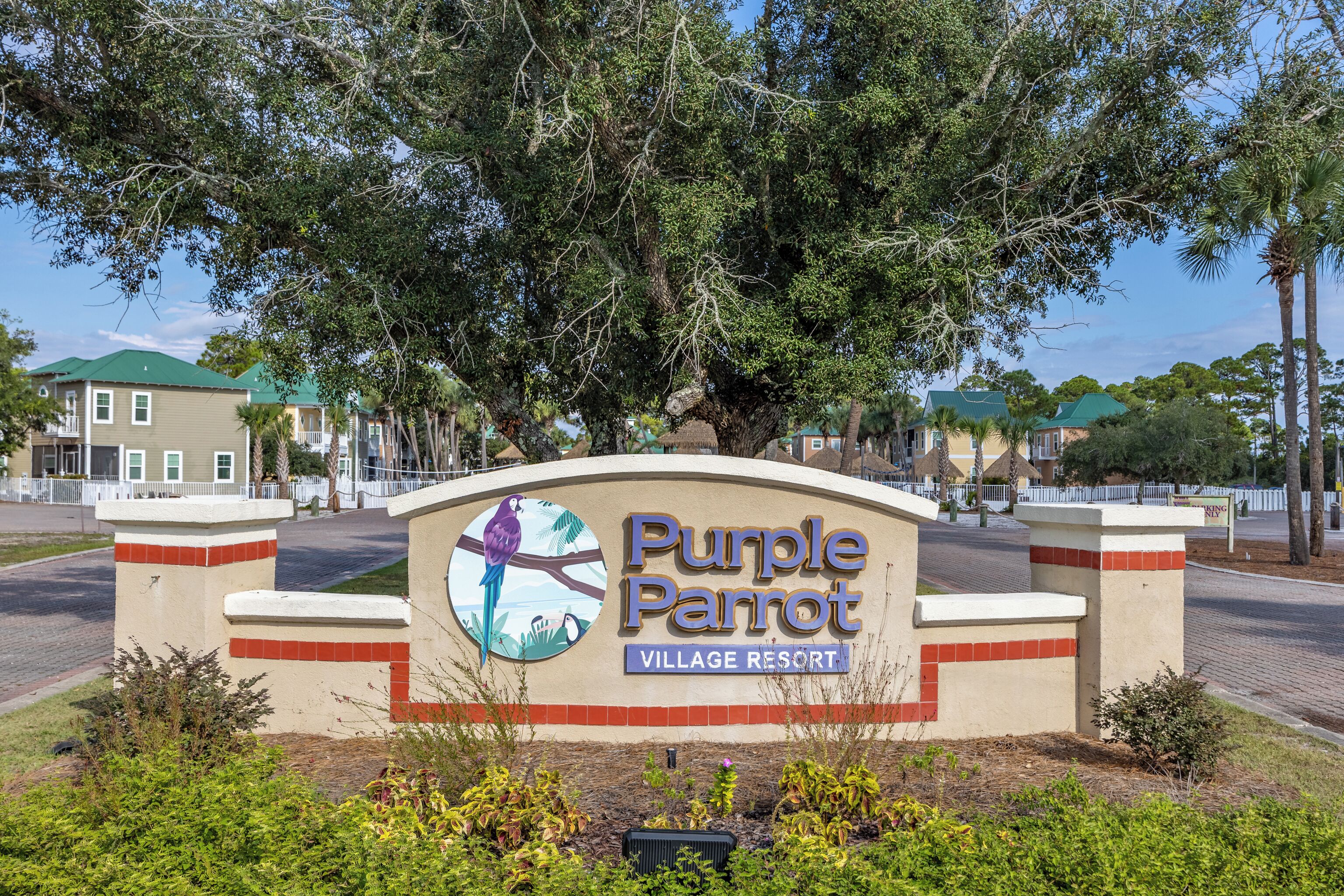 Placa do Purple Parrot Village Resort com o logotipo de um papagaio, rodeada por vegetação e edifícios.