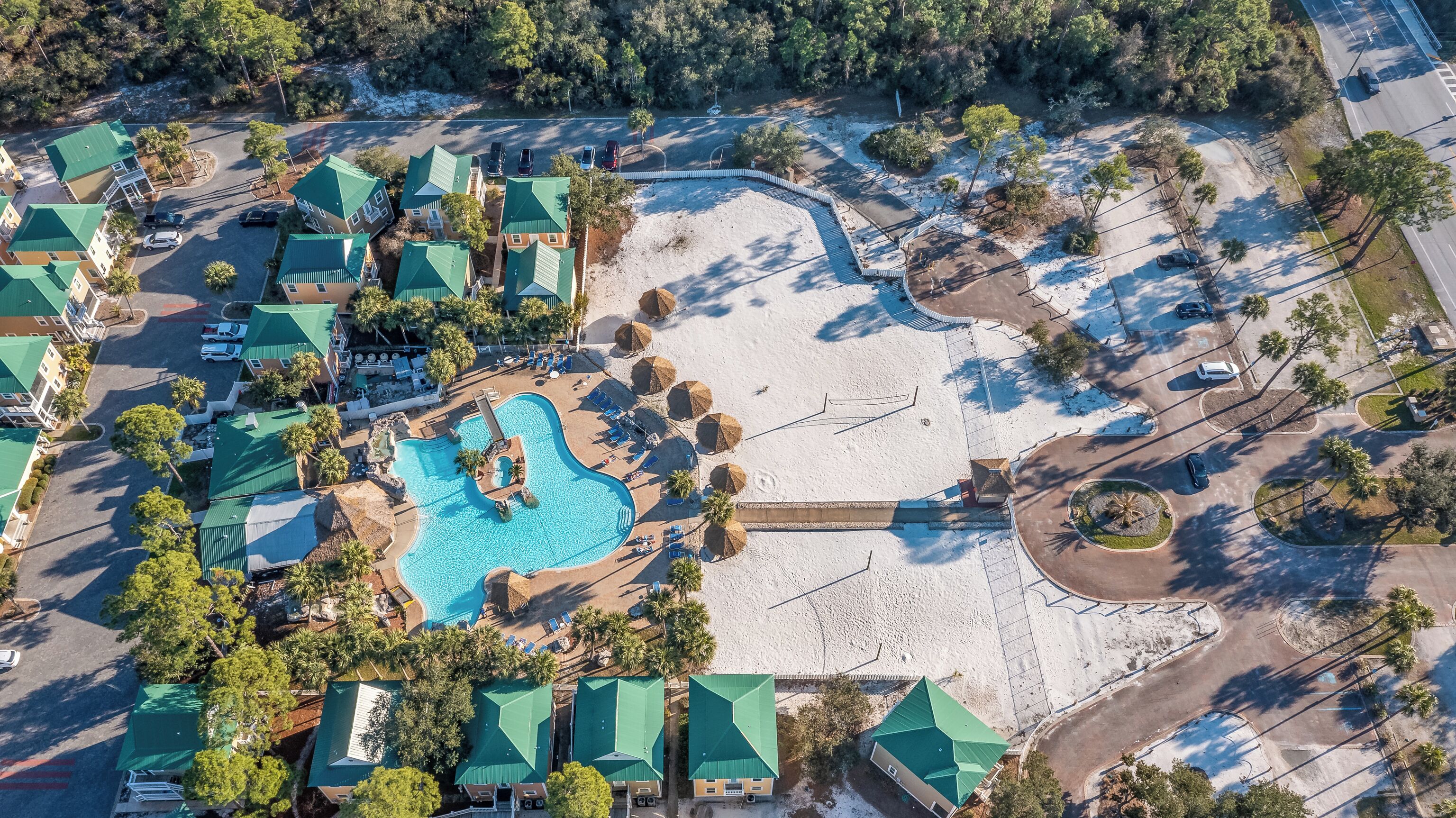 An aerial view of a resort with a central swimming pool, multiple buildings with green roofs, and a sandy beach area.