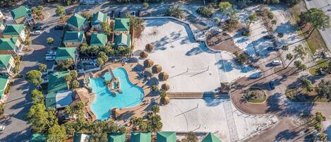 An aerial view of a resort with a central swimming pool, multiple buildings with green roofs, and a sandy beach area.