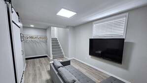 En moderne stue med en grå hjørnesofa, et fladskærms-tv monteret på væggen, en trappe til et andet niveau og et walk-in-closet med et tøjstativ.