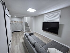 Ein modernes Wohnzimmer mit einem grauen Ecksofa, einem flachbildfernseher an der Wand, einer Treppe zu einer weiteren Ebene und einem begehbaren Kleiderschrank mit Kleiderstange.