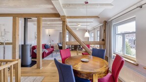 A modern interior with a round wooden table, red and blue chairs, a red sofa, and a large window providing a view of the outside. - 5* vacation home 188sqm with high-quality comfort with 4 bedrooms and space for 7 people (Sankt Andreasberg)