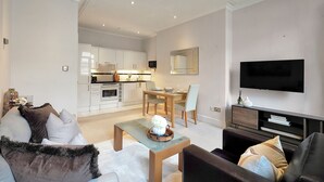A modern living room with a sofa, a coffee table, and a TV. There is a dining area with a table and chairs. The room has white walls and a kitchen in the background. - Hertford Flat 13 (London)