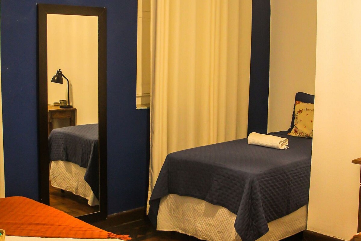A bedroom with a bed, a mirror, a bedside table with a lamp, a window with curtains, and a hanging light fixture.