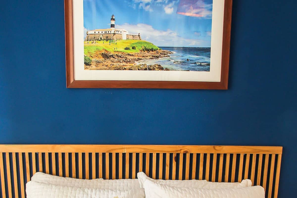 A neatly made bed with a wooden headboard, a framed picture of a lighthouse, and a basket with toiletries.