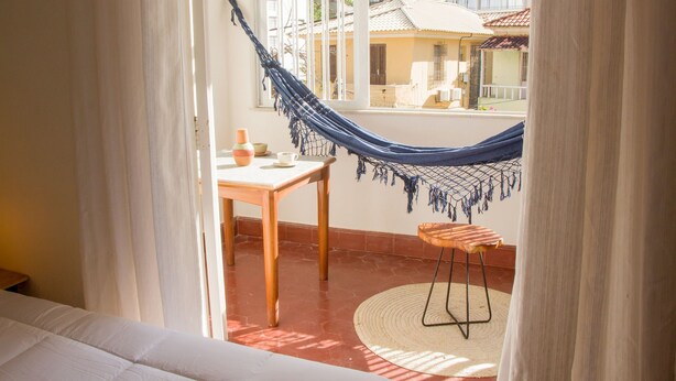 A room with a hammock, a wooden table, a small stool, and a view of buildings through a window. - Pousada Maria Maria (Salvador)