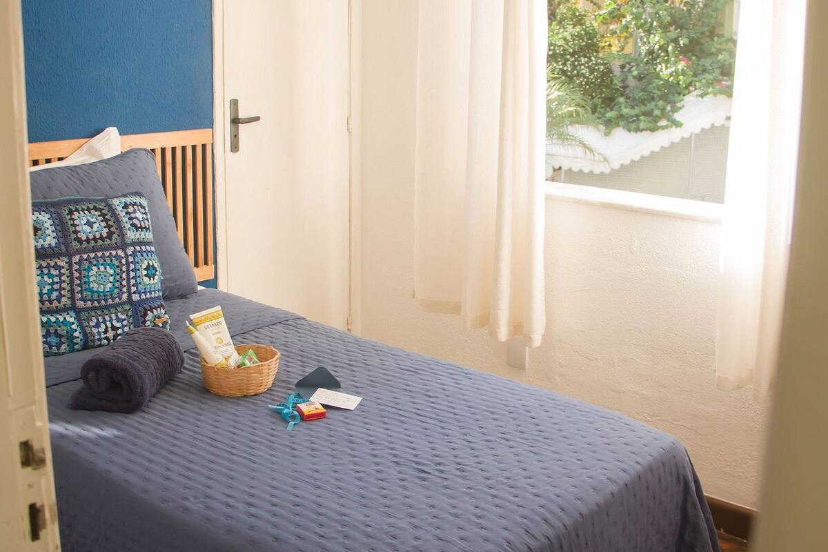 A bedroom with a bed, a window with curtains, a door, and a view of a building and greenery outside.