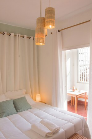 Une chambre à coucher avec un lit, des rideaux, un luminaire suspendu, une fenêtre et une table avec une chaise.