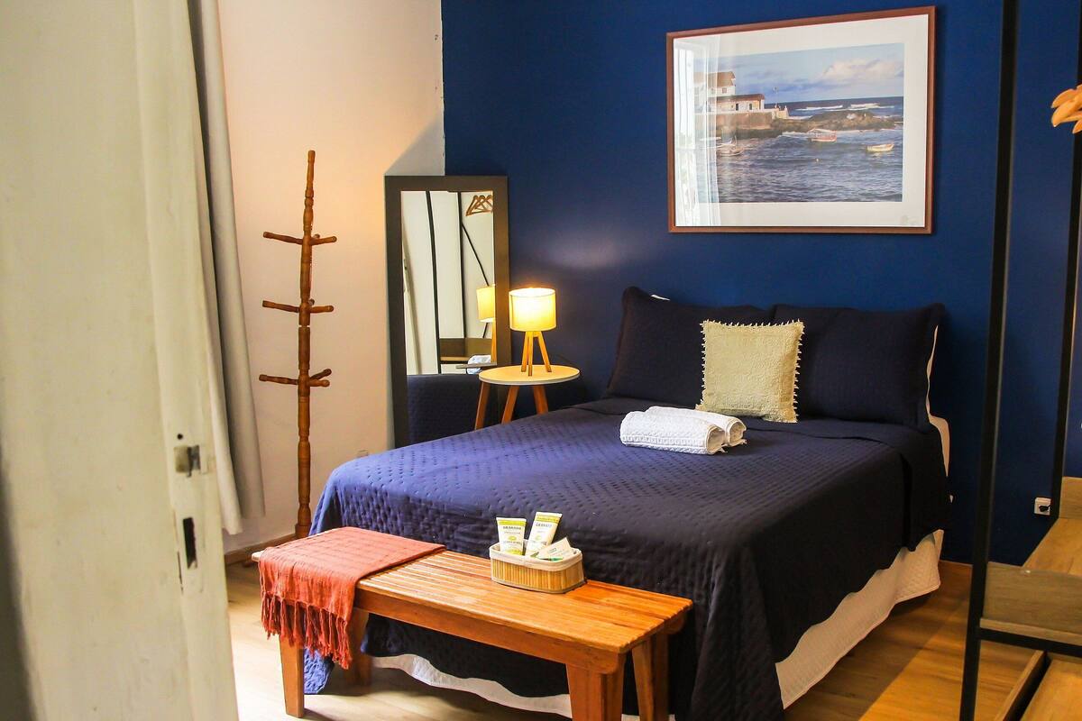A bedroom with a bed, bedside table, lamp, mirror, and a framed picture on the wall.