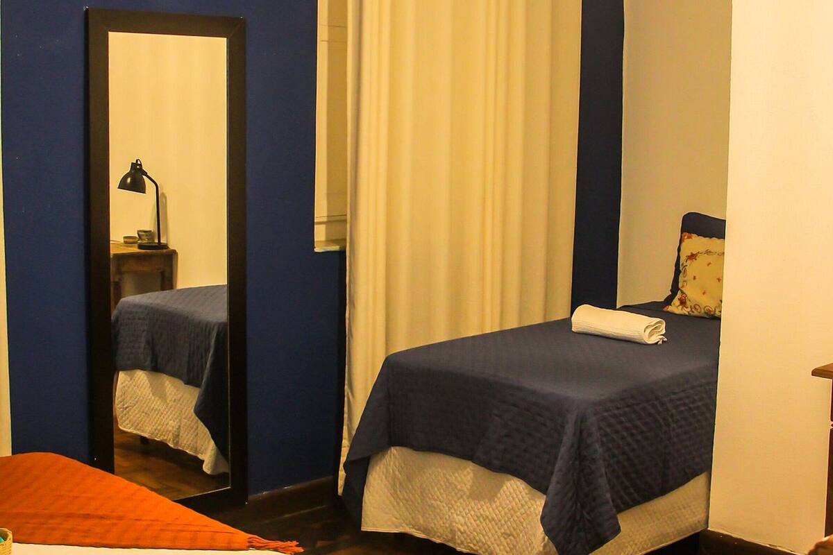 A bedroom with a bed, a mirror, a bedside table with a lamp, a window with curtains, and a hanging light fixture.