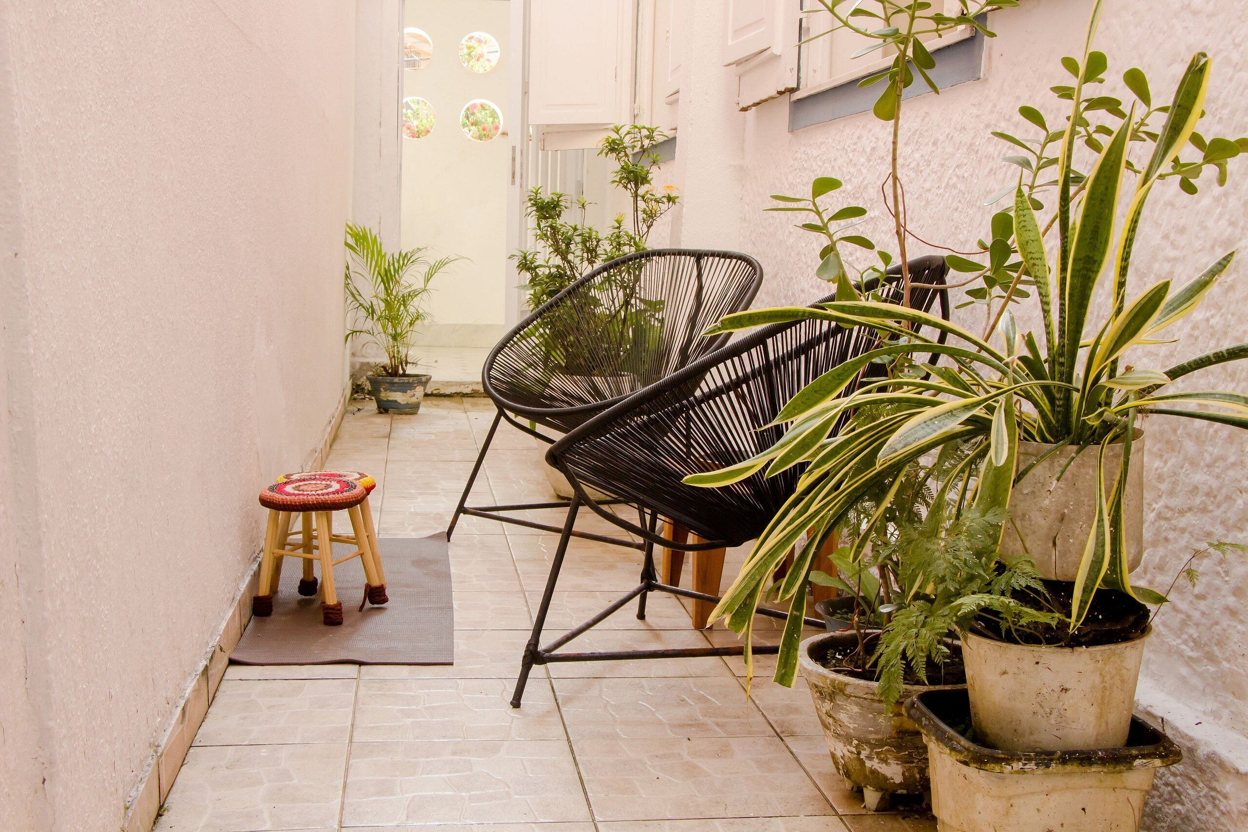 Um corredor exterior estreito com chão de azulejos, uma cadeira de metal preta, um banquinho de madeira e plantas em vasos.