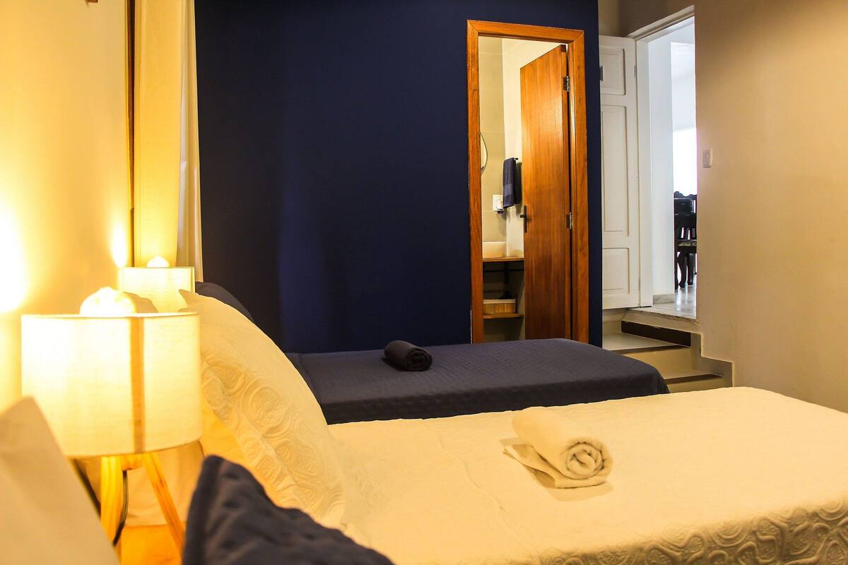 A hotel room with a bed, a bedside lamp, a small table, a mirror, and a door leading to a bathroom and another room.