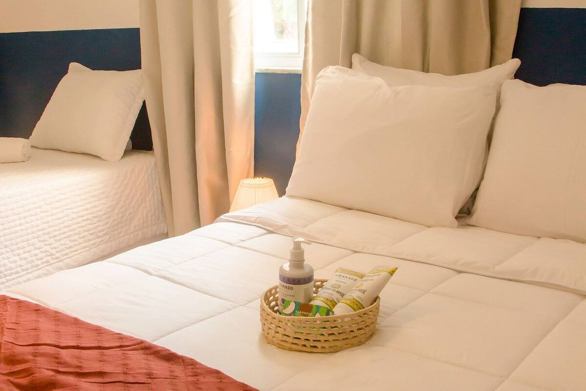 A hotel room with two beds, a bedside table with a lamp, and a basket with toiletries on the bed.
