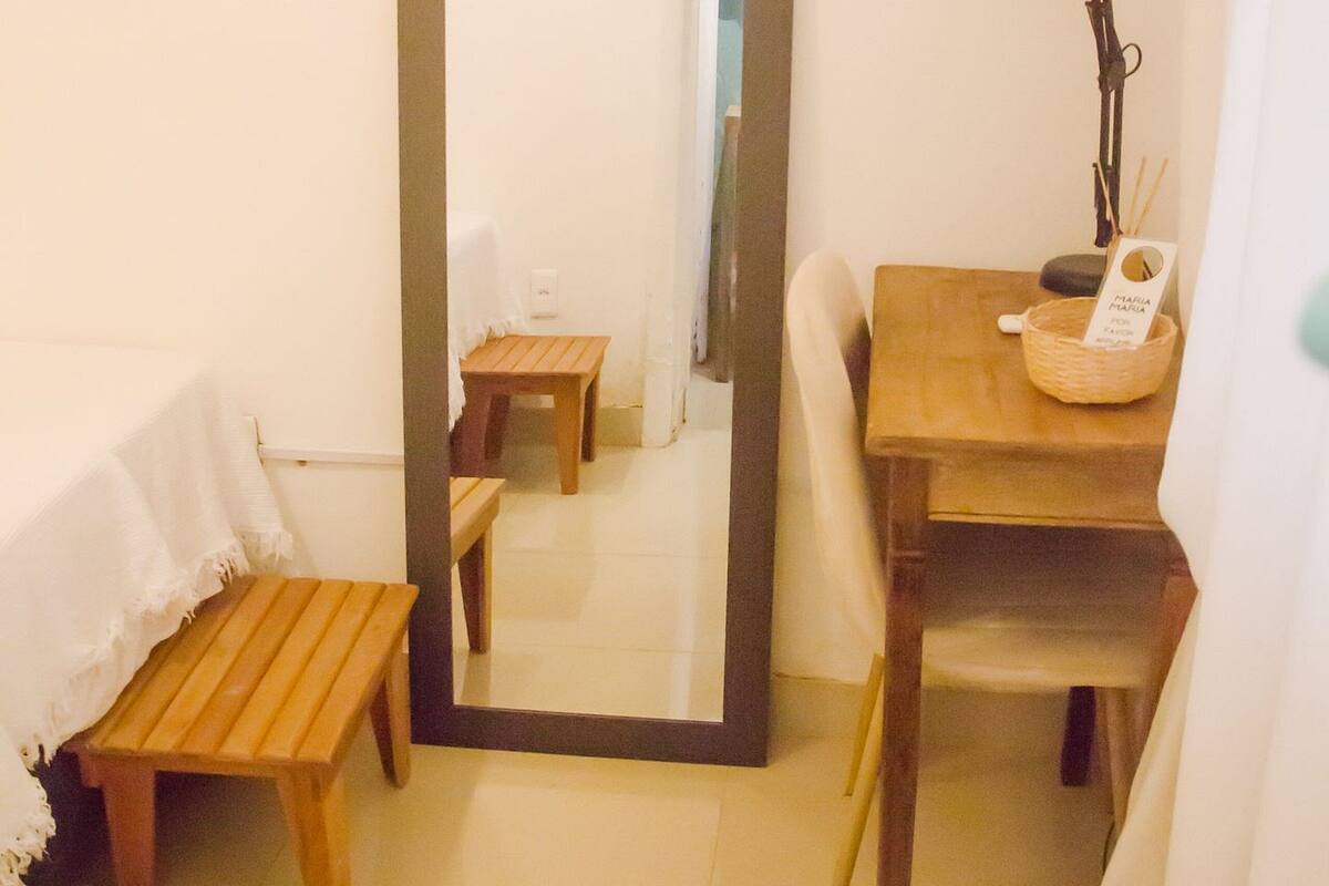 A small room with a wooden bench, a table with a lamp, and a mirror reflecting a door and a chair.