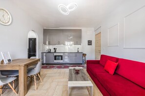 A modern living room with a red sofa, a wooden dining table, and a kitchen area with grey cabinets and a white countertop. - Saranda Bay Retreat - 1BR Apartment (Sarandë)