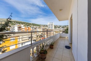 A balcony with a view of a hillside town, potted plants, and a metal railing. - Saranda Bay Retreat - 1BR Apartment (Sarandë)