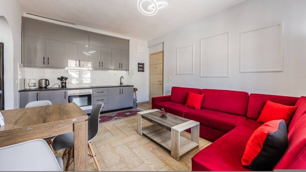 A modern living room with a red sofa, a wooden coffee table, and a kitchen area with grey cabinets and a white countertop. - Saranda Bay Retreat - 1BR Apartment (Sarandë)