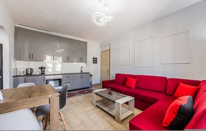 A modern living room with a red sofa, a wooden coffee table, and a kitchen area with grey cabinets and a white countertop. - Saranda Bay Retreat - 1BR Apartment (Sarandë)