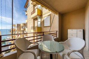 Ein Balkon mit Korbmöbeln und einem Glastisch, mit Blick auf eine Küstenlandschaft und andere Gebäude.