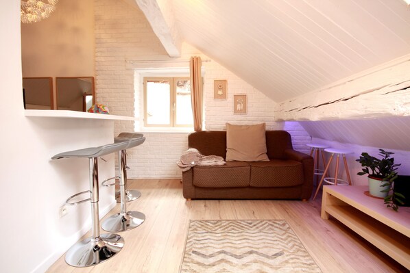 Un acogedor espacio tipo loft con suelo de madera, un sofá marrón, una pequeña mesa y una zona de bar con taburetes.