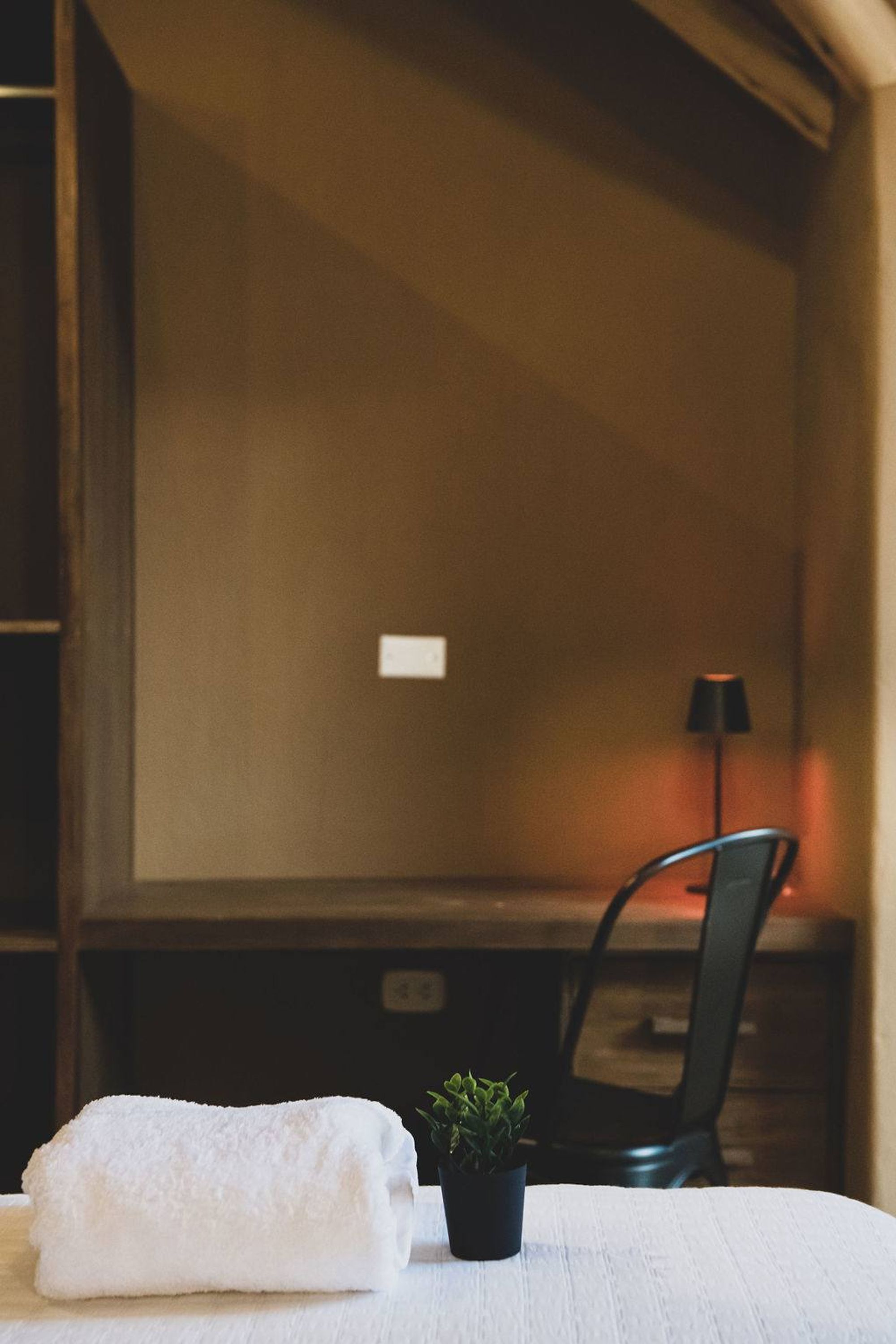 A neatly made bed with a white towel, a small potted plant, and a chair in a room with wooden walls and a lamp.