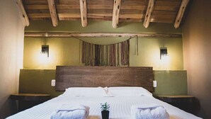 Hotellihuone, jossa on puinen katto, suuri sänky valkoisilla vuodevaatteilla, kaksi yöpöytää ja pieni kasvi sängyn päällä.