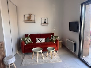 Un salon moderne avec un canapé rouge, deux petits tabourets, un tabouret blanc à poils longs et une porte en verre donnant sur l’extérieur.
